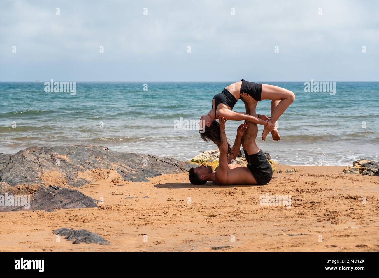 Side view of woman in black activewear lying on raised legs of man and stretching back during acro yoga session on sandy beach near waving sea Stock Photo