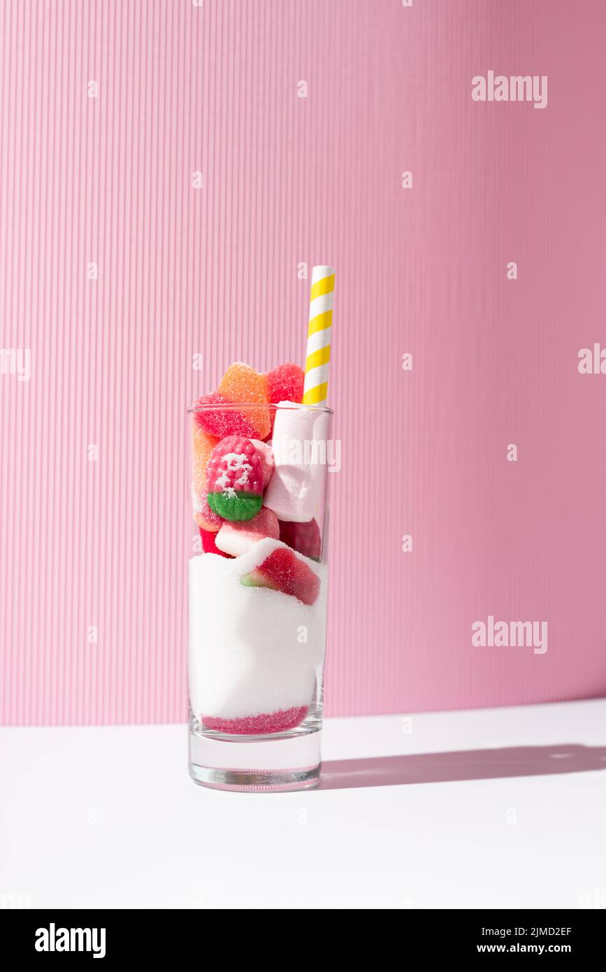 Glass with bunch of sweet gummy candies and white sugar placed near striped straw on pink and white background Stock Photo