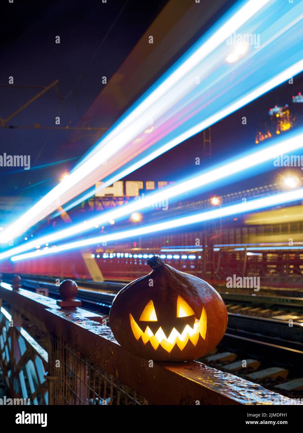 Halloween pumpkin with a glowing grimace at night on the railway bridge. In the background there are rails and blurry streaks of Stock Photo