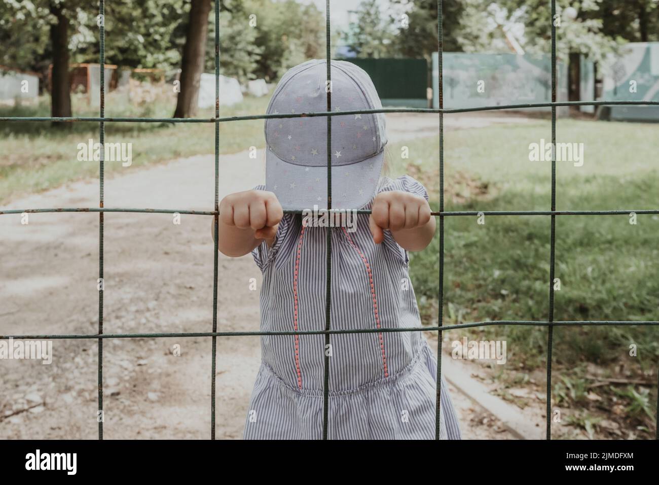 Little baby girl behind bars. The child holds onto the bars of the lattice. Stock Photo