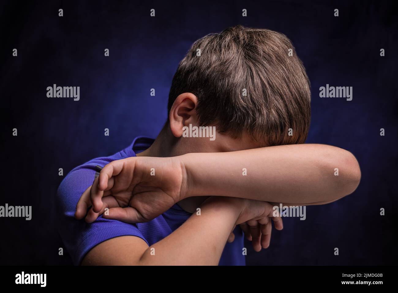 Sad european teenage boy with light brown hair in sports purple t-shirt covering his face with one hand over isolated background Stock Photo