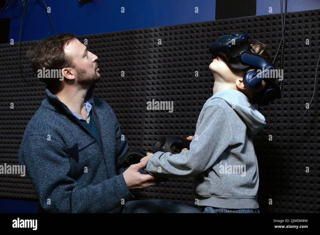 Man helps boy in virtual reality helmet with joysticks in his hands understand how to control video game Stock Photo