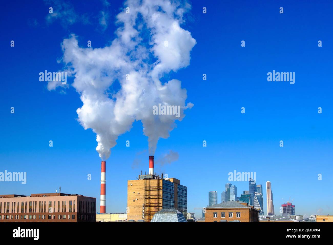 White puffs of smoke rise up from the industrial chimneys above the city in the blue cloudless sky. Stock Photo