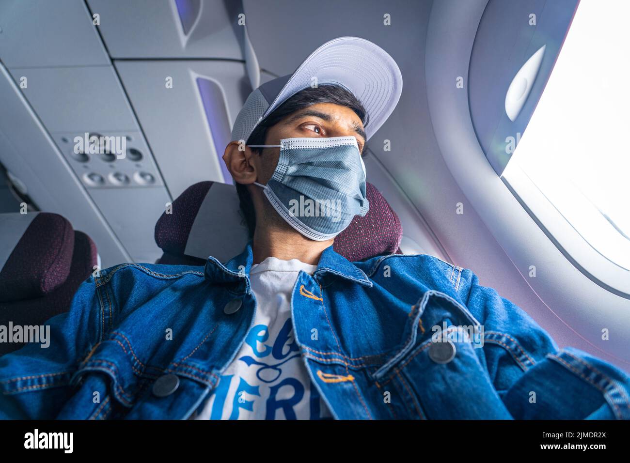 A young adult traveler wearing a mask and a grey hat looks outside the airplane window. Stock Photo