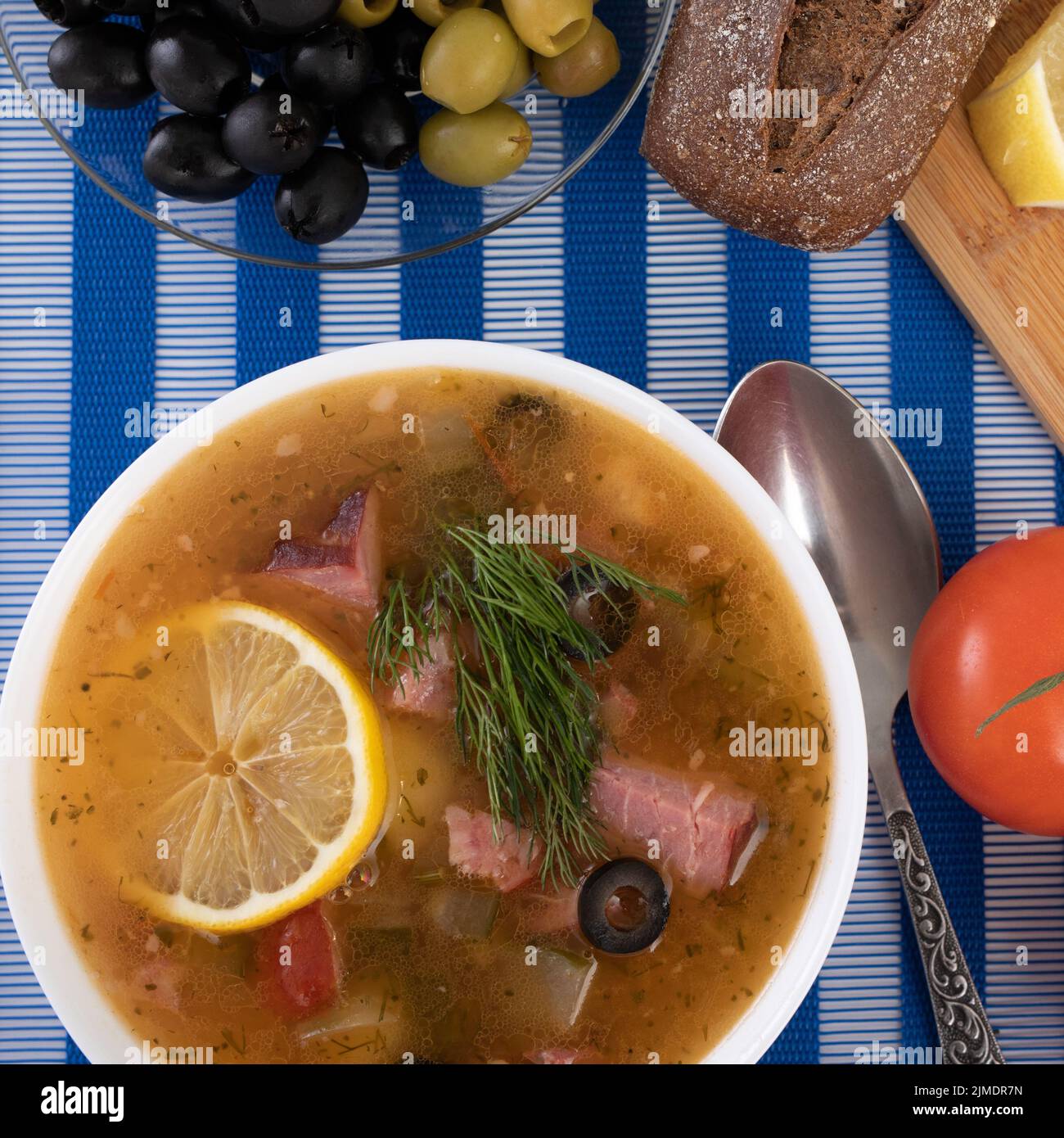 Traditional Russian soup Solyanka cooked with meat, sausages, salted cucumbers on wite bowl. Black bread, lemon, olived and toma Stock Photo