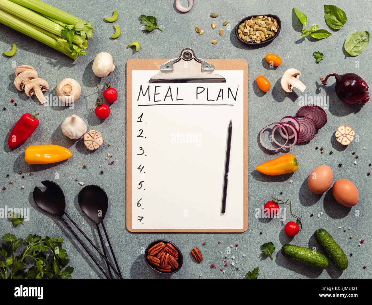 Meal plan concept. Food ingredients, salad serving utensils and clipboard with letters MEAL PLAN and seven numbers. Gray background. Diet menu concept Stock Photo