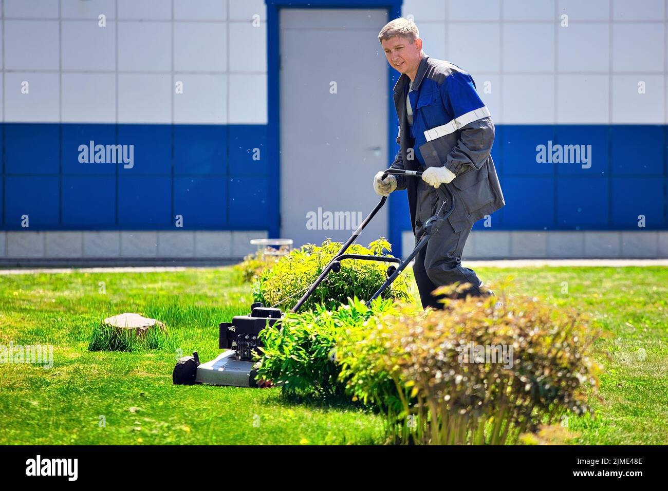 A young man walks with a lawn mower on a green lawn in a production area. Stock Photo