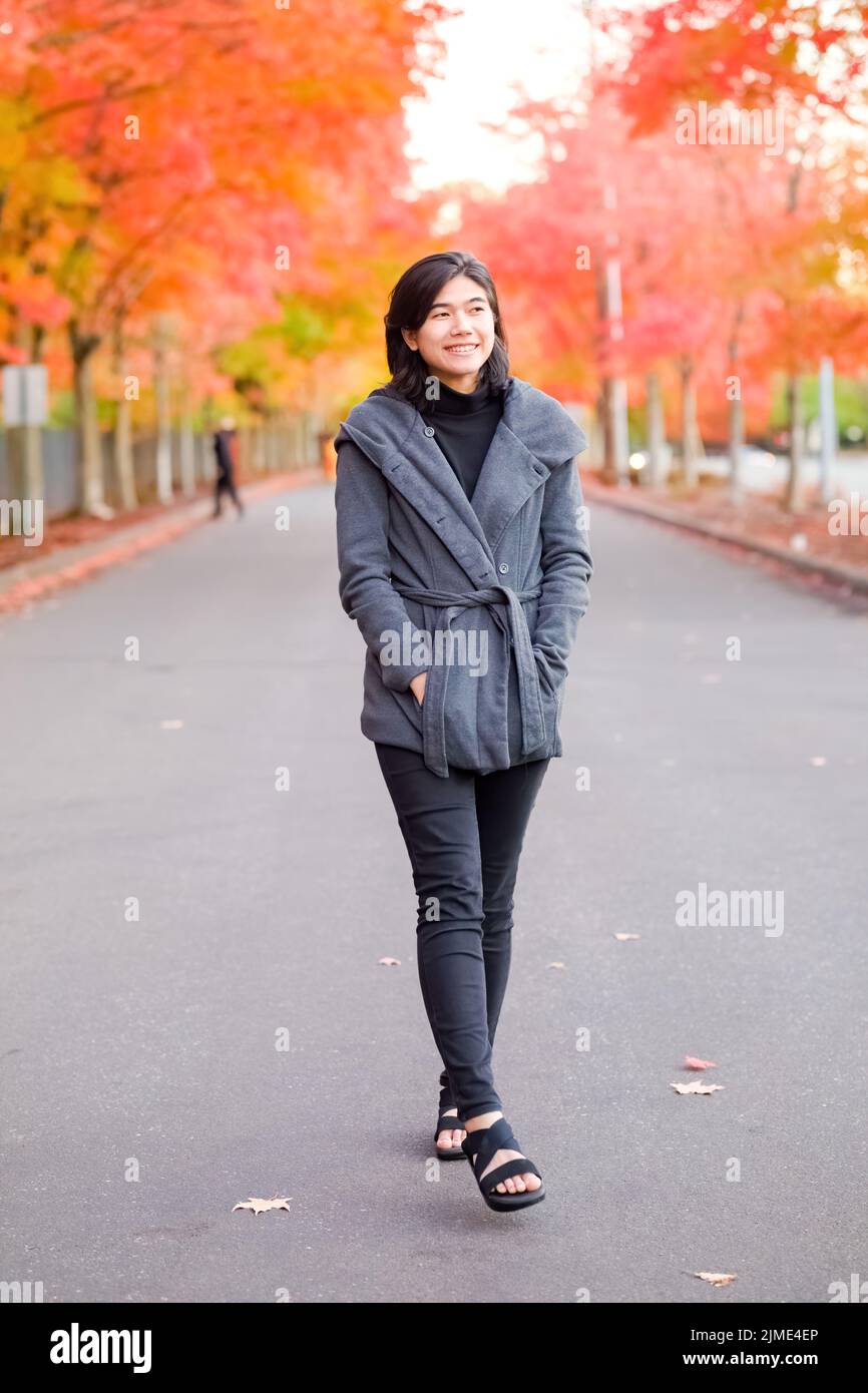 Smiling biracial teen girl or young adult female in gray jacket walking along road enjoying colorful autumn leaves Stock Photo