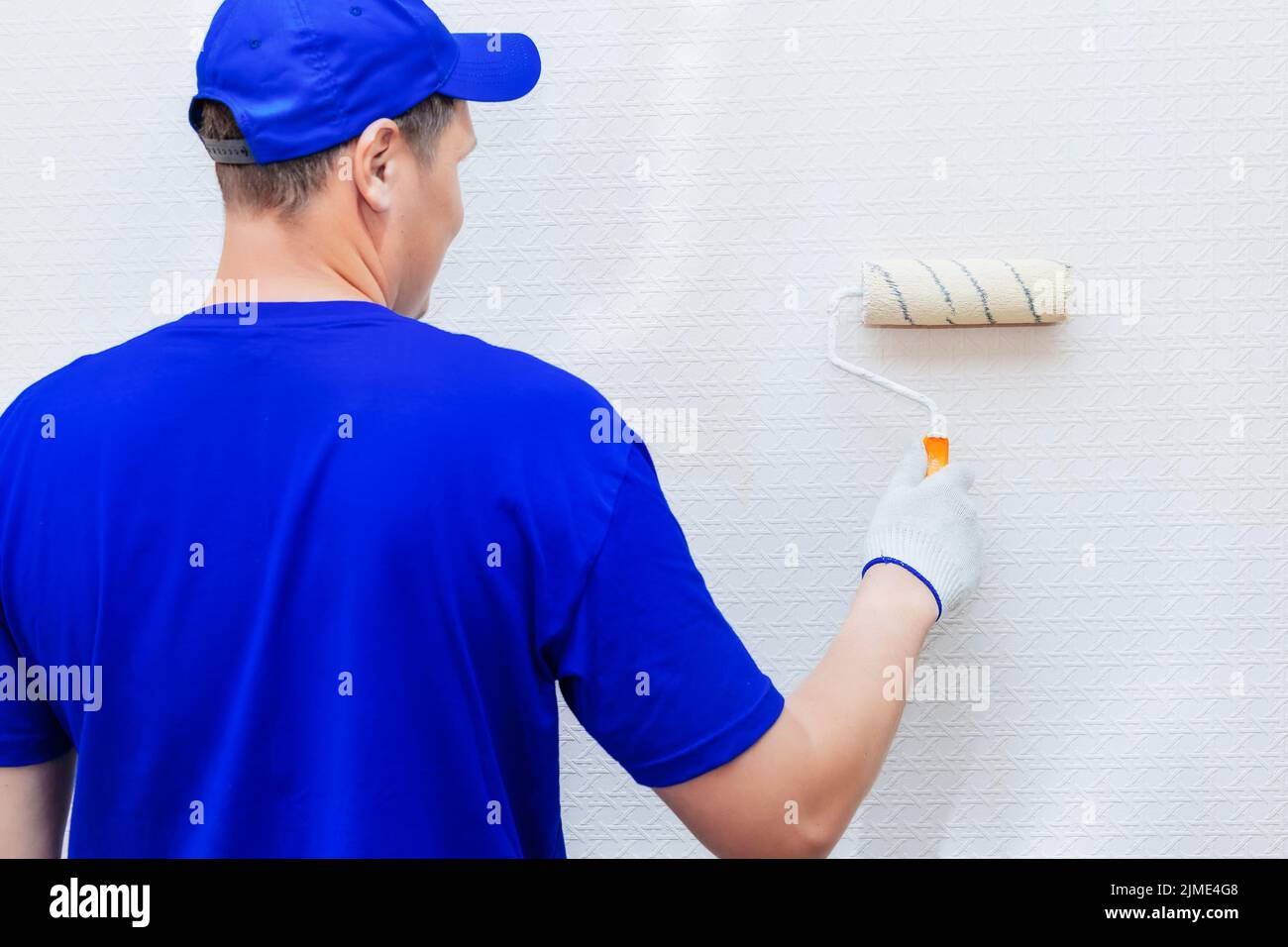 A painter in a cap and gloves paints the wall with a paint roller. Portrait of a young worker in uniform. Stock Photo