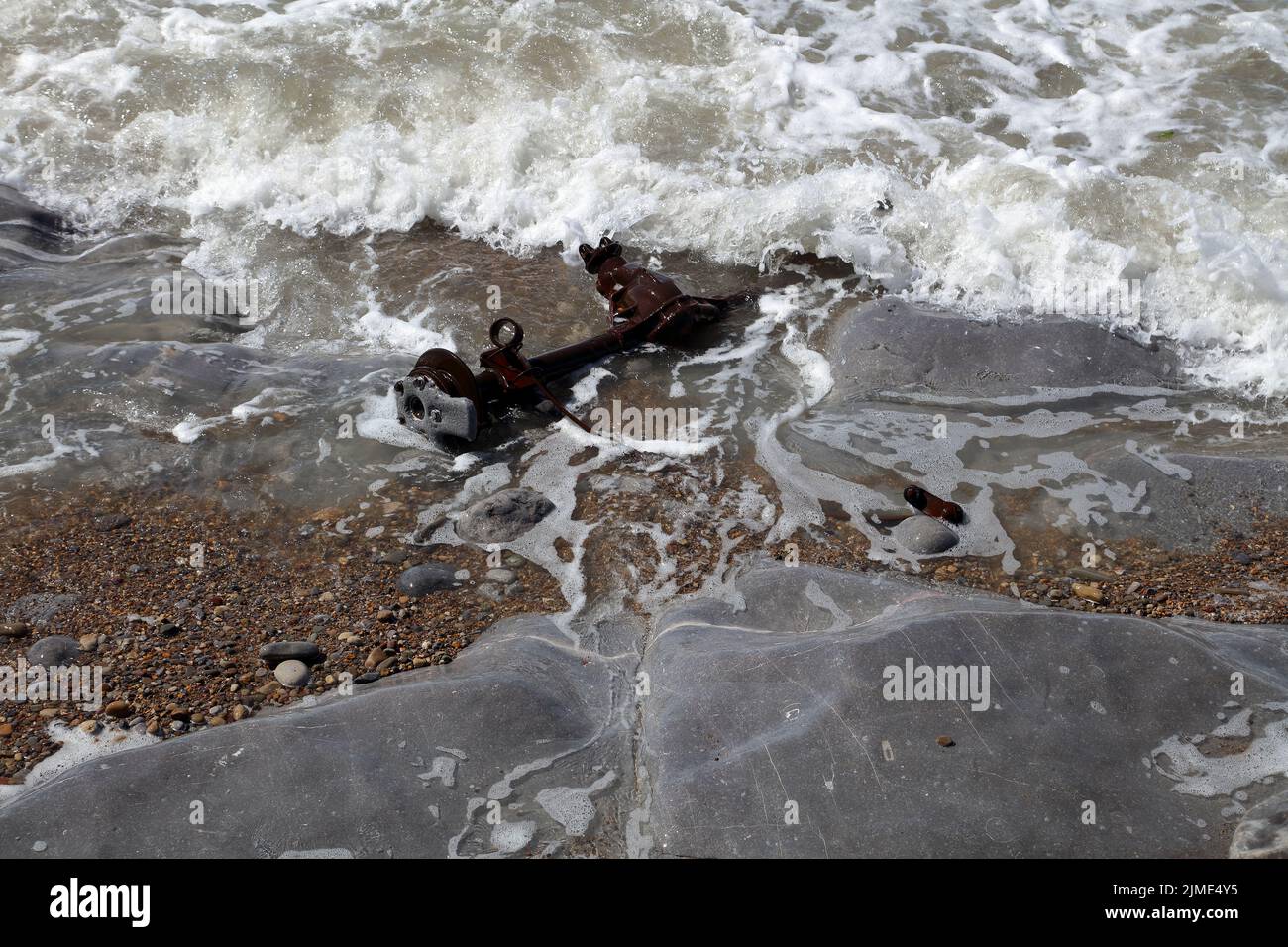 The mangled rear axle of a car stranded on the beach being washed over by the incoming tide. Stock Photo