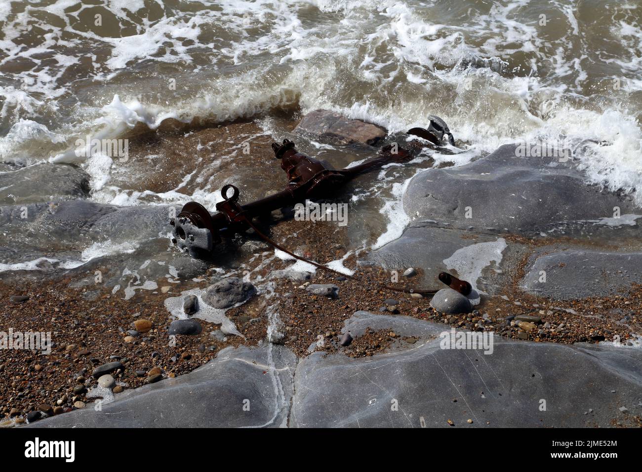 The mangled rear axle of a car stranded on the beach being washed over by the incoming tide. Stock Photo