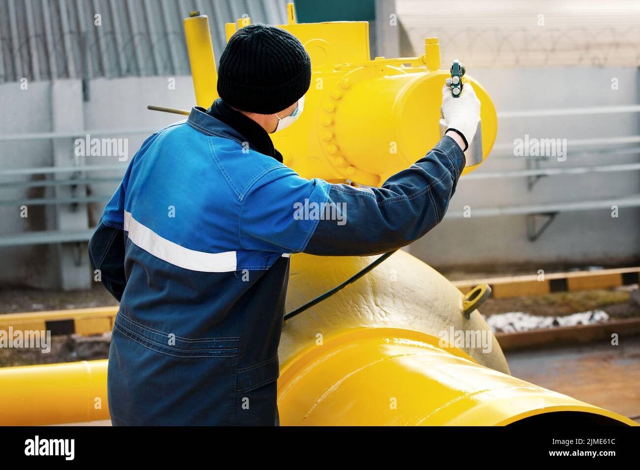 A painter in working clothes paints a metal shut-off valve for gasification from a compressor gun on a summer day. Stock Photo