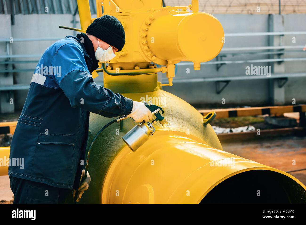 A painter in working clothes paints a metal shut-off valve for gasification from a compressor gun on a summer day. Stock Photo