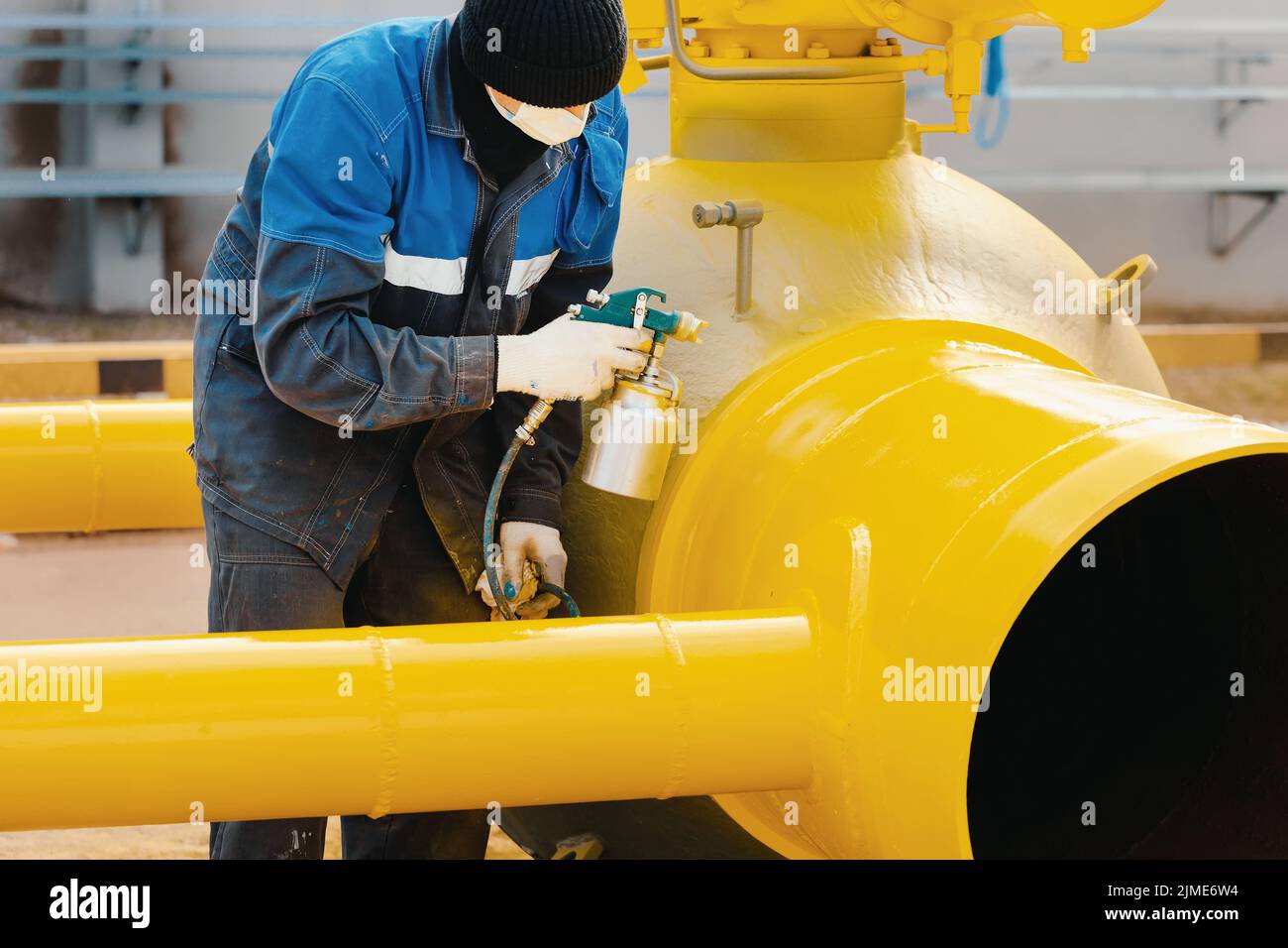 A painter in working clothes paints a metal shut-off valve for gasification from a compressor gun on a summer day. Stock Photo