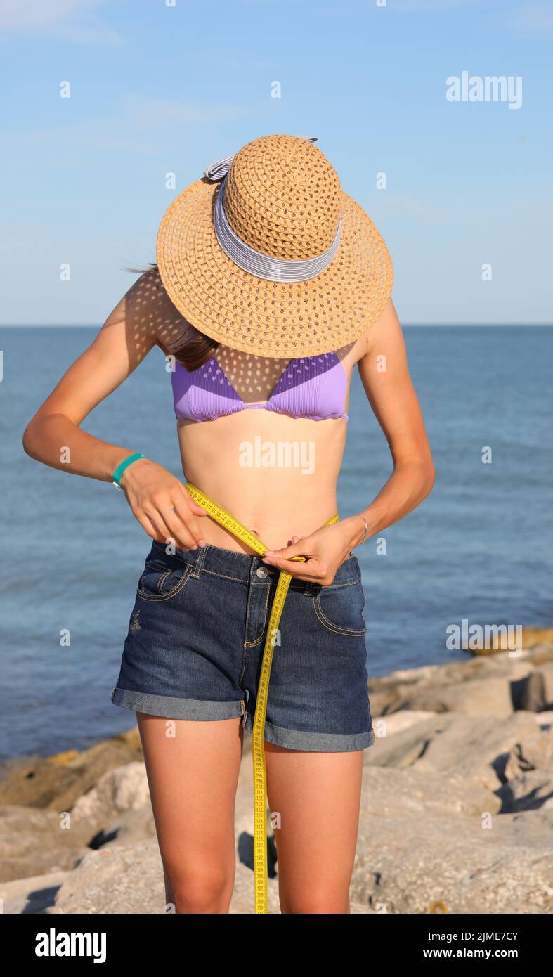 young skinny girl in bikini with straw hat while measuring her waist with a flexible tape measure Stock Photo