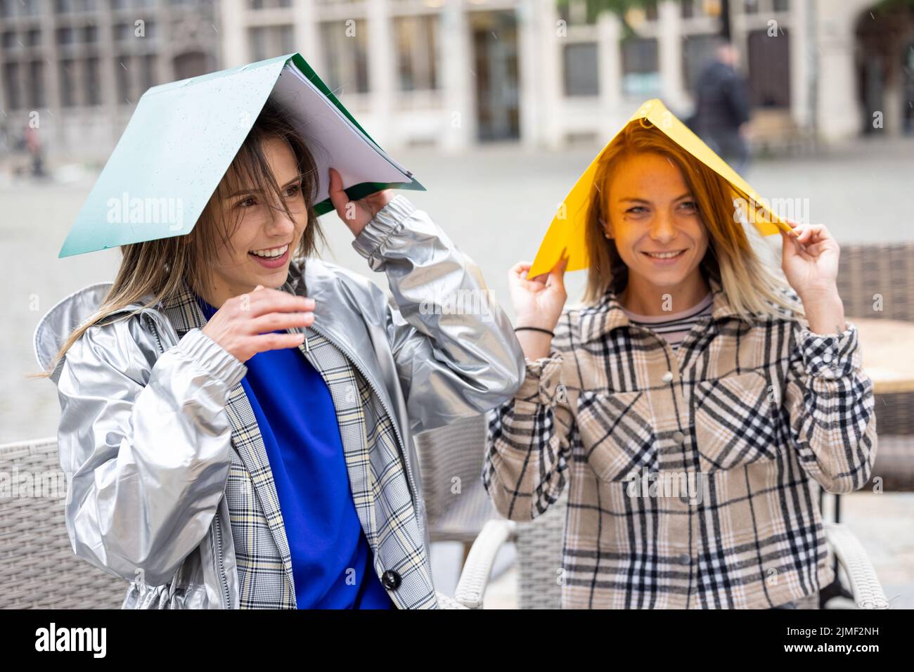 Two female students sitting outside at a terrace table covering their heads for the rain with two colored plastic folders Stock Photo