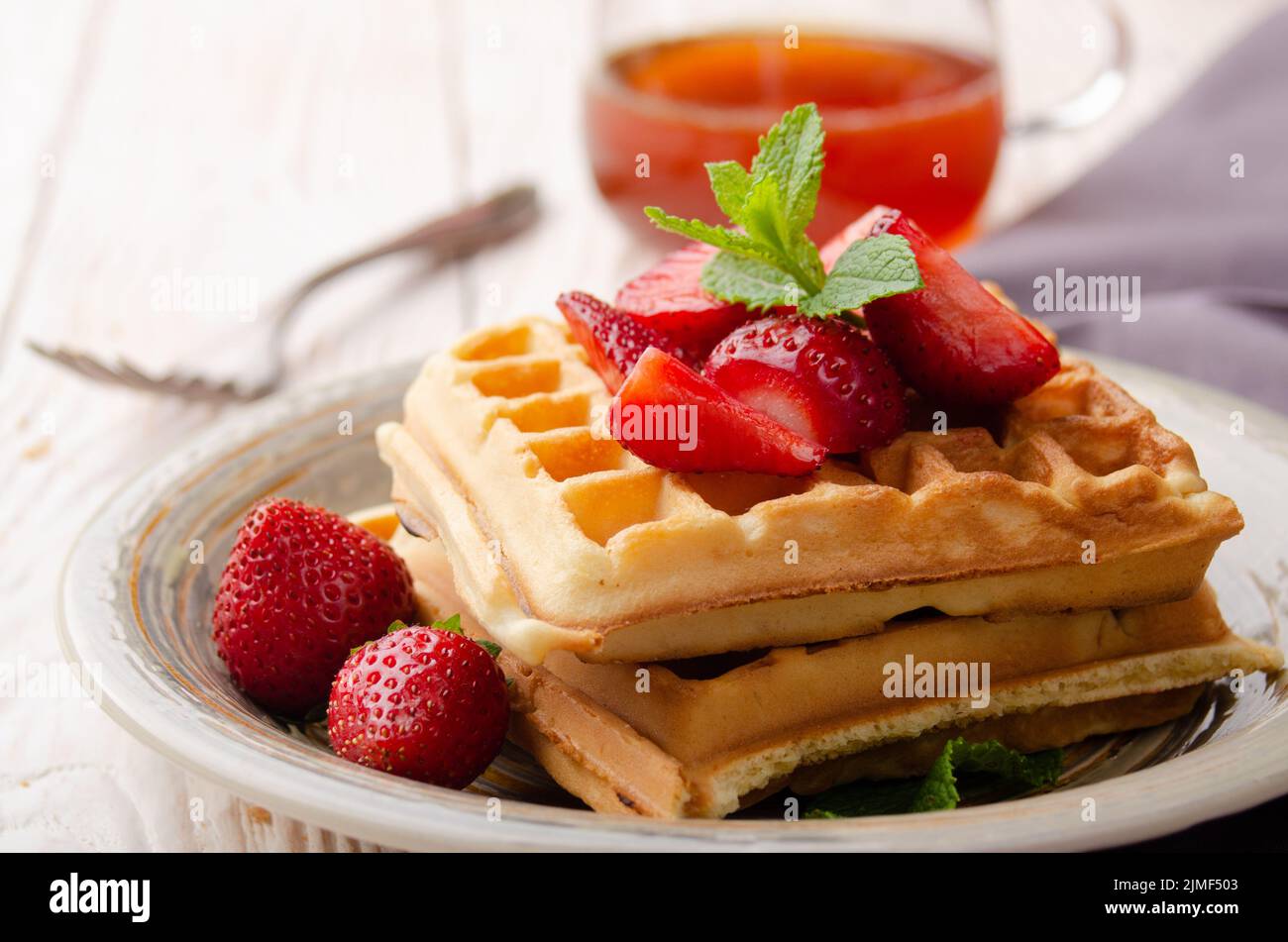Closeup view at belgian waffles served with strawberries and mint leaf on white wooden kitchen table with syrup aside Stock Photo