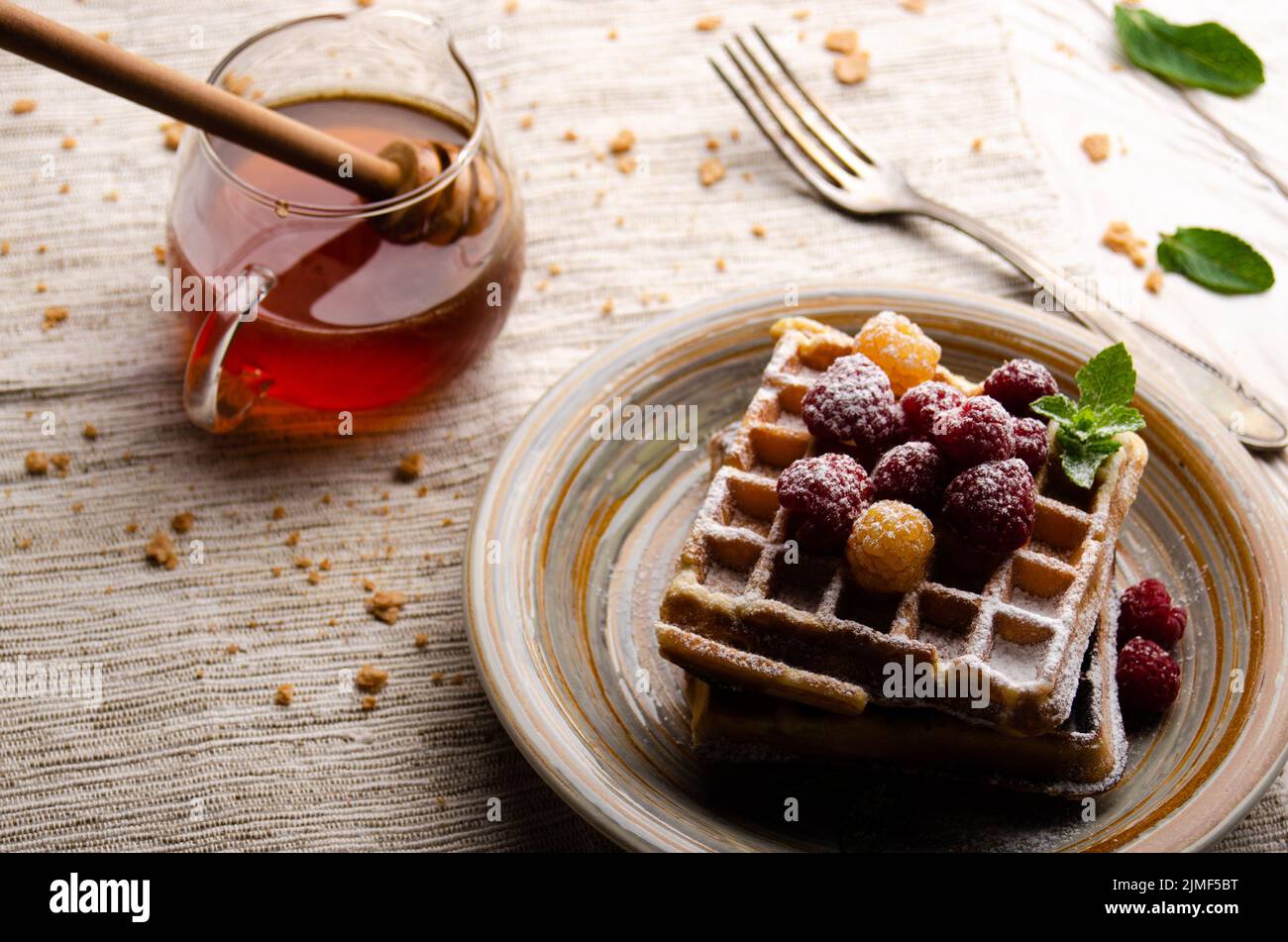 Belgian waffles served with raspberries and mint leaf dusted with powdered sugar on white wooden kitchen table with syrup aside. Stock Photo