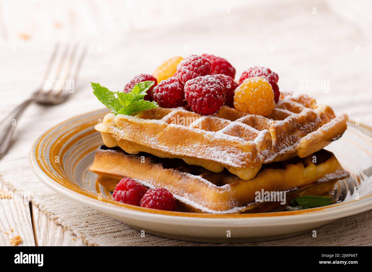 Belgian waffles served with raspberries and mint leaf dusted with powdered sugar on white wooden kitchen table Stock Photo