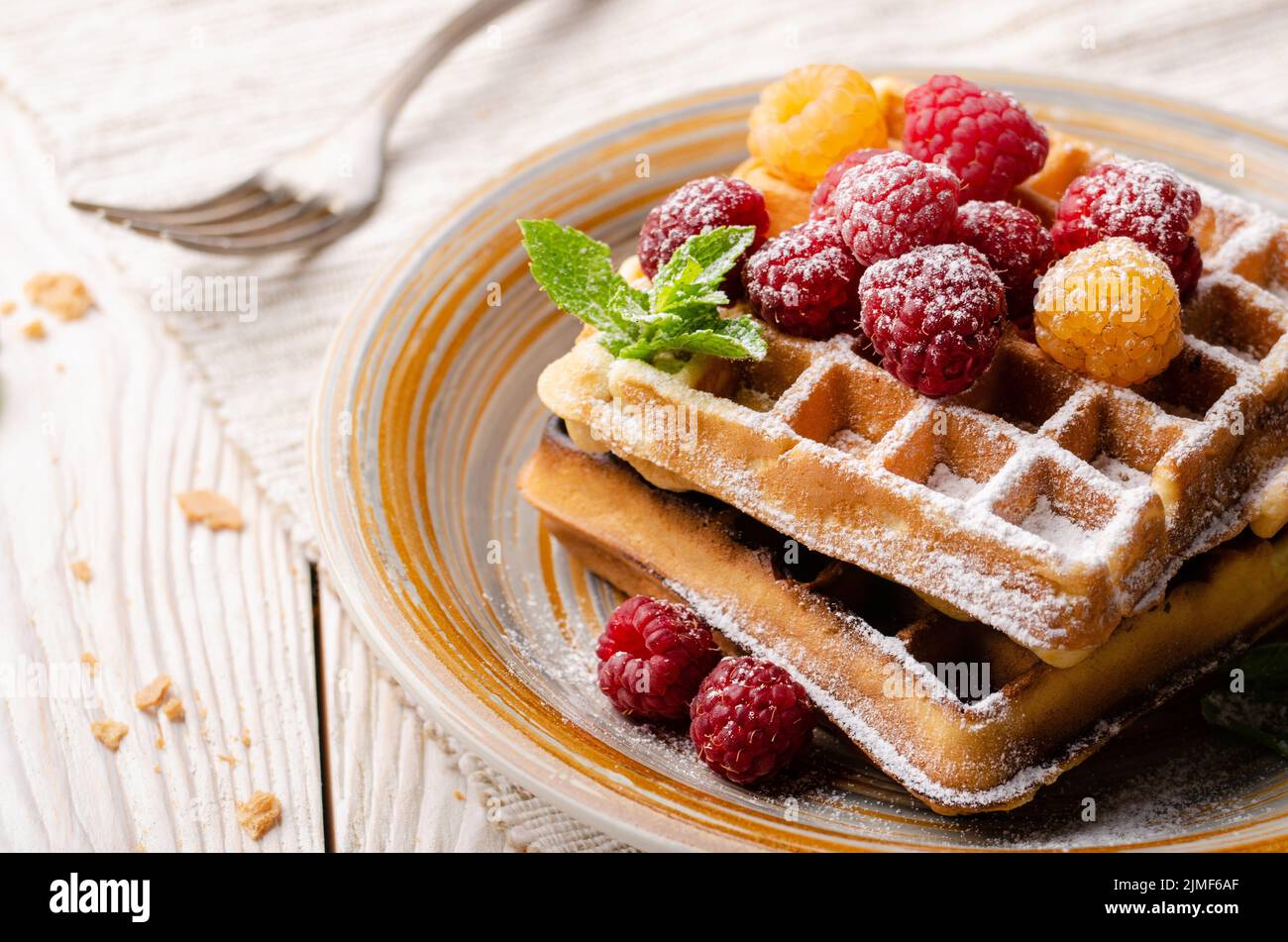 Belgian waffles served with raspberries and mint leaf dusted with powdered sugar on white wooden kitchen table Stock Photo