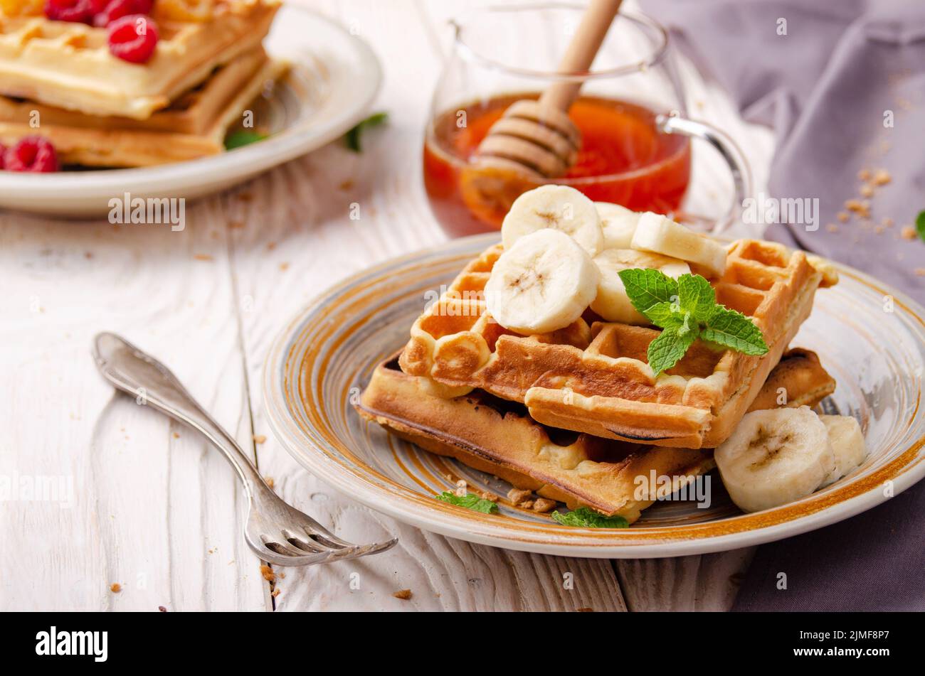 Belgian waffles served with banana and mint leaf on white wooden kitchen table with syrup aside Stock Photo