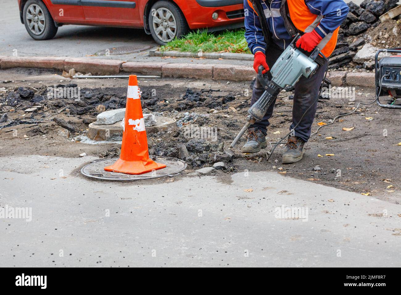 A worker using an electric jackhammer and a gasoline generator cleans the area near the sewer manholes from the old asphalt. Stock Photo