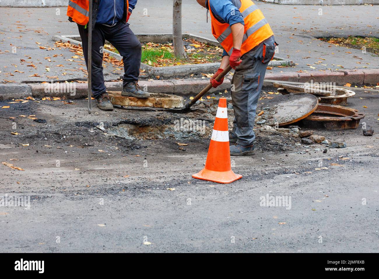 A team of road workers with a shovel and a crowbar clean a place for a new sewer manhole on the roadway on a summer day. Copy space. Stock Photo