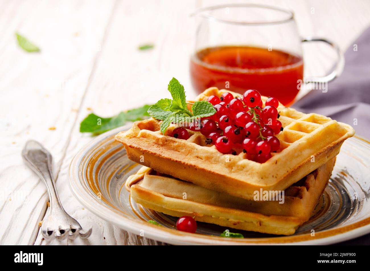 Belgian waffles served with red currants and mint leaf on white wooden kitchen table with syrup aside Stock Photo