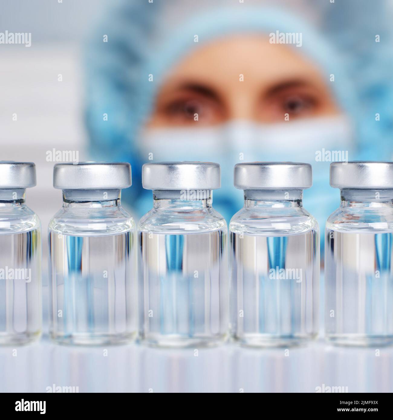 Female nurse in safety mask and cap looks at vials with liquid medicine on refrigerator shelf Stock Photo