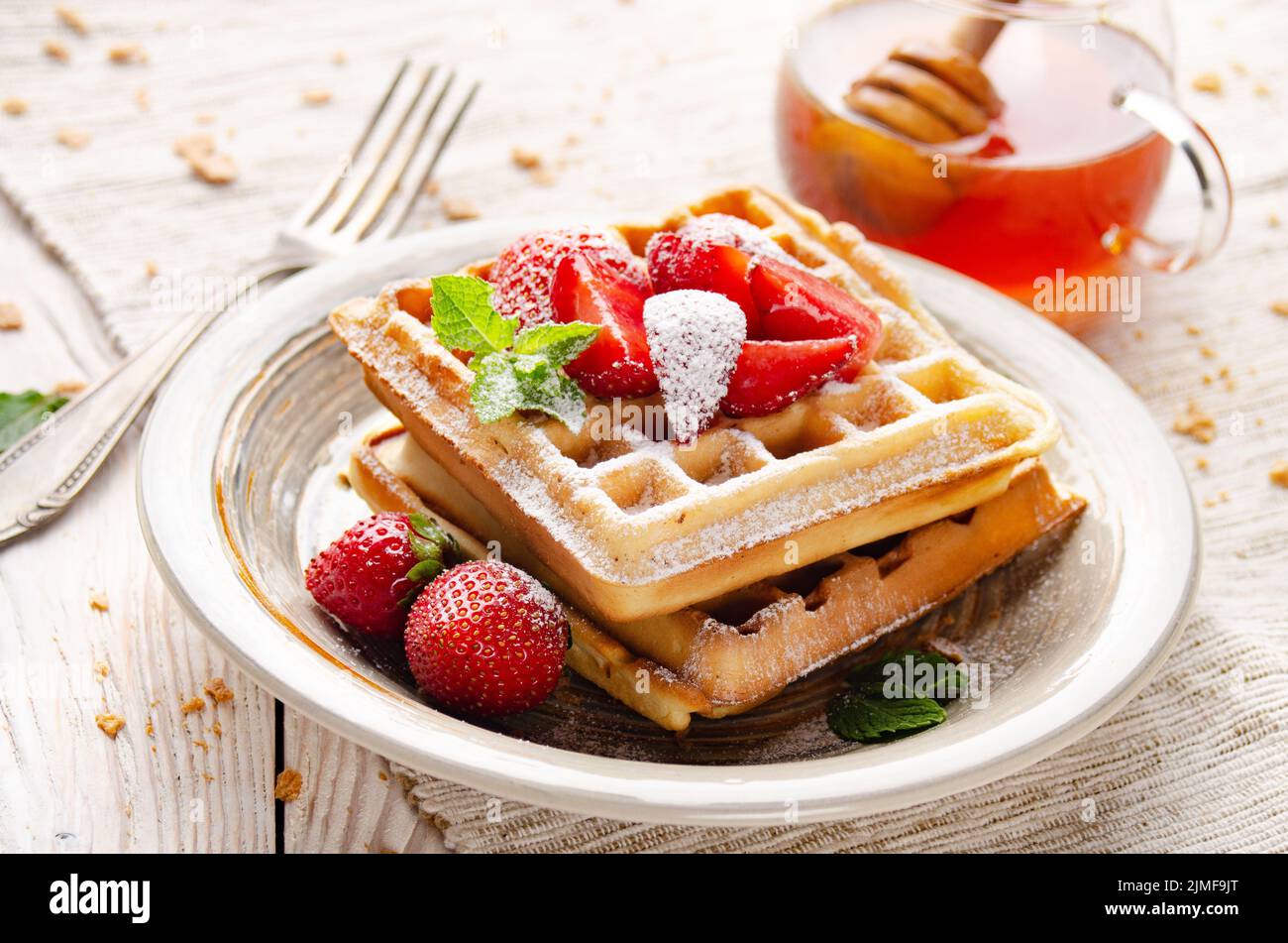 Belgian waffles served with strawberries and mint leaf dusted with powdered sugar on white wooden kitchen table with syrup aside Stock Photo