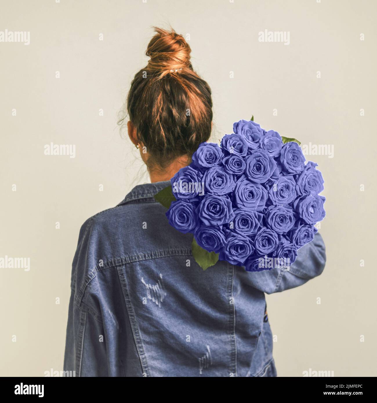 Back view of young woman in denim jacket holding bunch of violet roses on shoulder. Girl with bun updo in jeans holding flowers in very peri color 202 Stock Photo