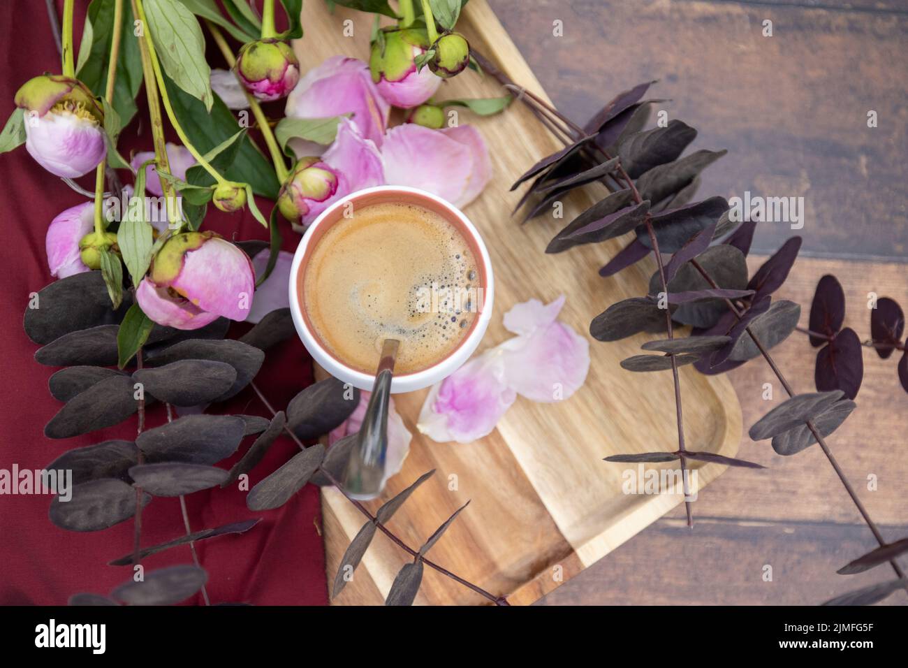 A white Cup of black coffee stands on a wooden background next to pink peonies and other garden flowers and leaves, The concept Stock Photo