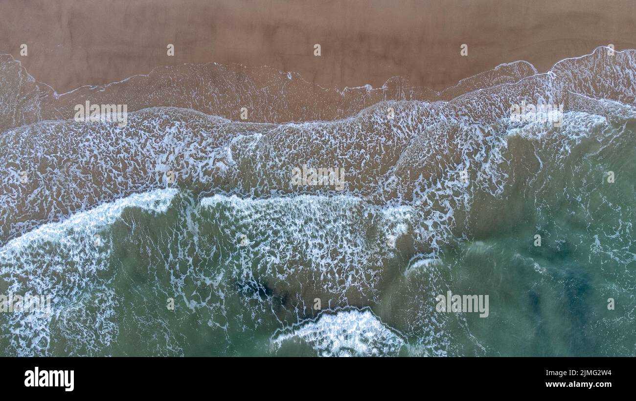 Aerial drone view perpendicular to the blue waves crashing on the sandy beach at the coast. The impact with wet wax creates whit Stock Photo