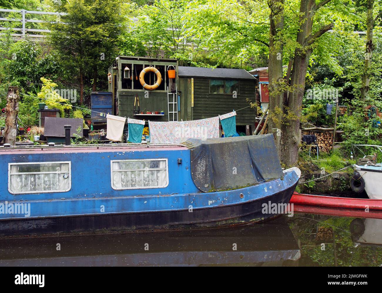 An old scruffy blue narrow boat moored next to wooden sheds with washing on the line on the rochdale canal near hebden bridge Stock Photo