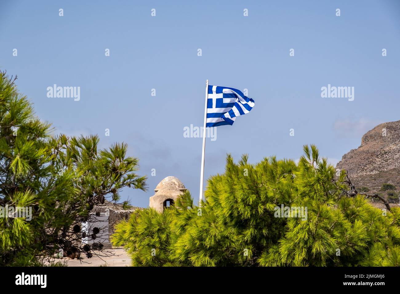 Greek flag waves in front of old building at Venetian Castle or Fortezza, monument at Kythira Ionian islands, Greece. View over green tree, blue sky b Stock Photo