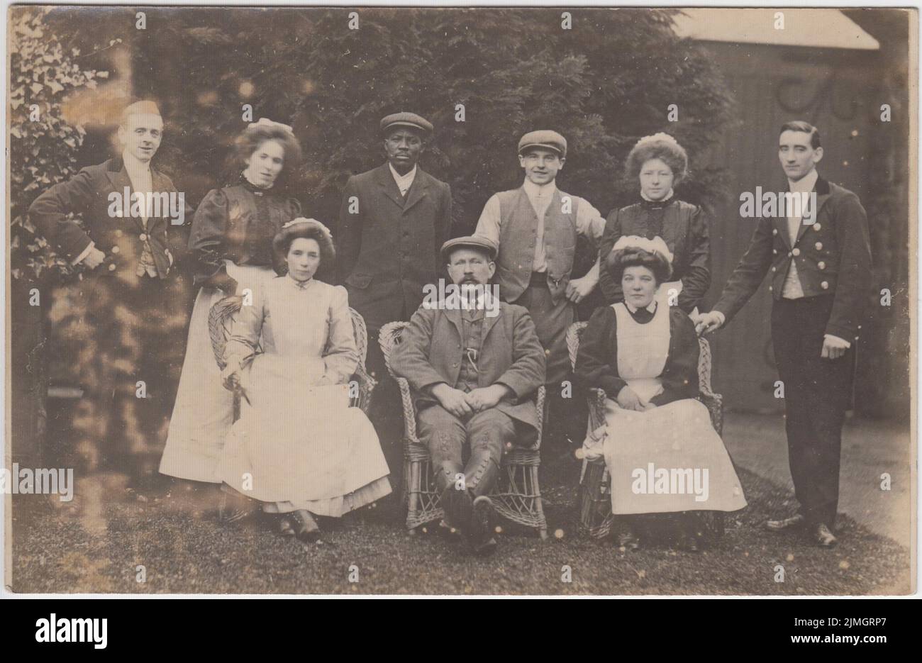 Early 20th century photograph of a group of servants posing for the camera in a garden. The servants include four maids in uniforms, two men in smart buttoned jackets (footmen?), and three men in suits and flat caps. One of the men is African - Caribbean or Black British Stock Photo