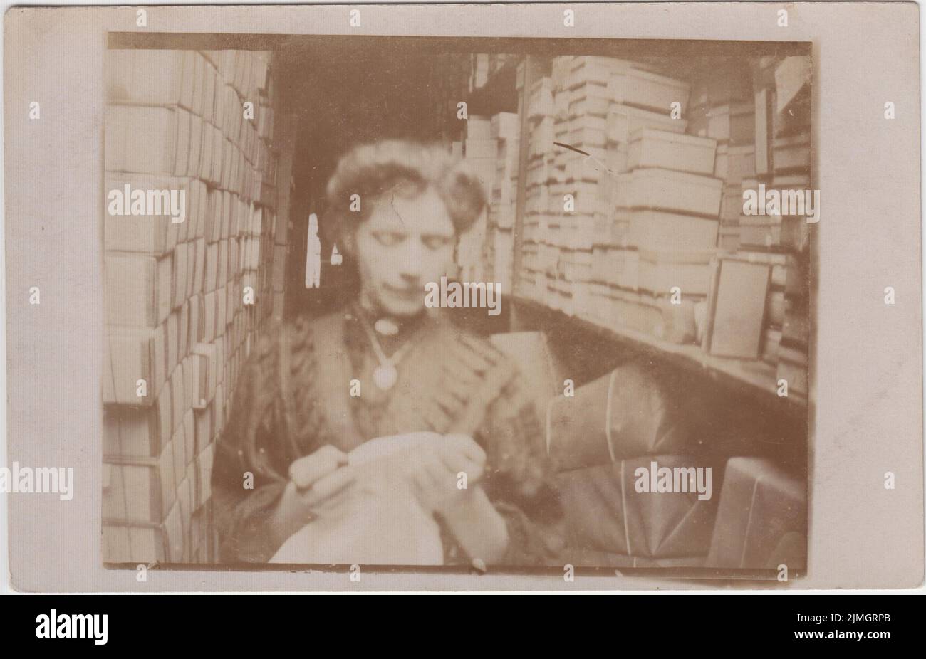 Woman photographed in the storeroom of a shop or warehouse in the Hackney area of London, early 20th century. She is examining a piece of fabric, and boxes and parcels tied up with brown paper and string are on the shelves behind her Stock Photo