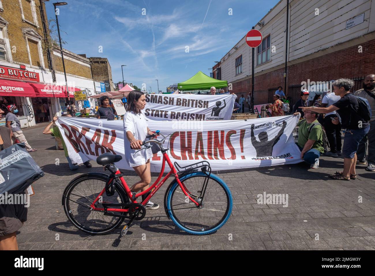 London, UK. 6 Aug 2022. A woman wheels her bike past the 'Break The Chains' banner of the Revolutionary Communist Group who spoke, handed out leaflets and collected petition signatures in front of busy Ridley Road market opposite Dalston Kingsland station. They say the UK government policies are clearly racist, exploiting migrant workers who come here and creating a hostile environment with immigration raids, treating the Windrush families disgracefully, organising charter deportation flights and planning to send refugees to Rwanda. Peter Marshall/Alamy Live News Stock Photo