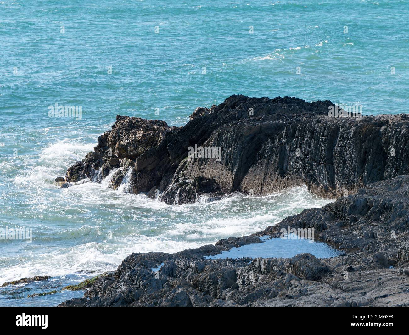 Tidal waves break on black coastal rocks, seascape. The water is turquoise in color. White foam on the waves, splashes, rock beside sea. Stock Photo