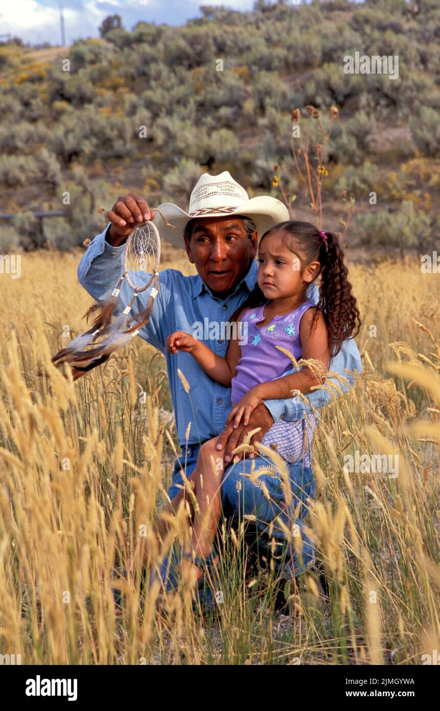 Native American family of a grandfather with his granddaughter sitting on his knee in a field as he tells her the dreamcatcher story Stock Photo