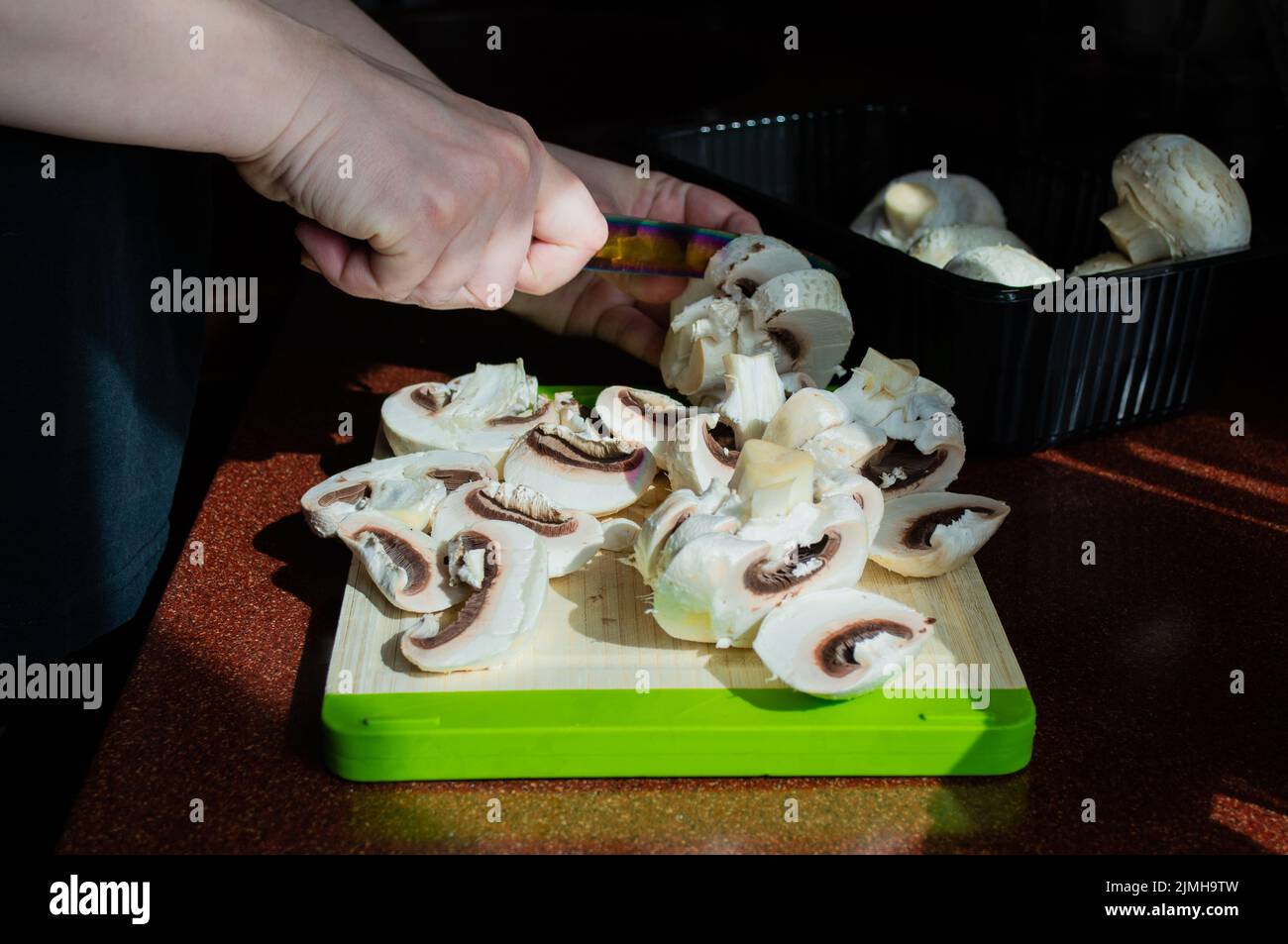 Close-up of hands with sliced fresh mushrooms on a cutting board. Homemade vegetarian cuisine. Sunlight from window, shadows on the table. Stock Photo