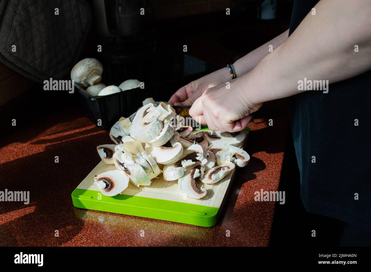 Close-up of hands with sliced fresh mushrooms on a cutting board. Homemade vegetarian cuisine. Sunlight from window, shadows on the table. Stock Photo