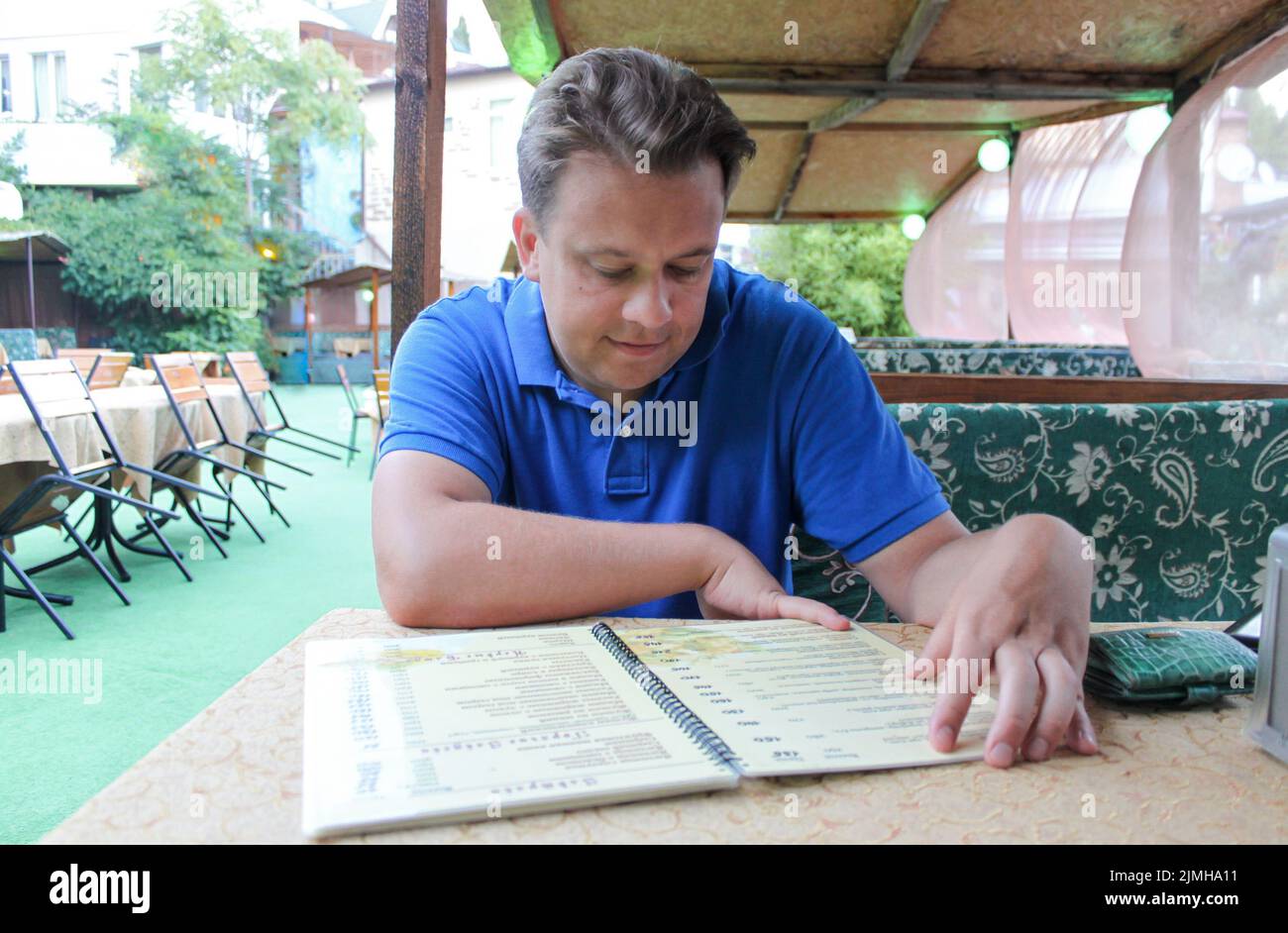 A young man on veranda of a summer restaurant looks at the menu. He is clearly confused by prices. Stock Photo