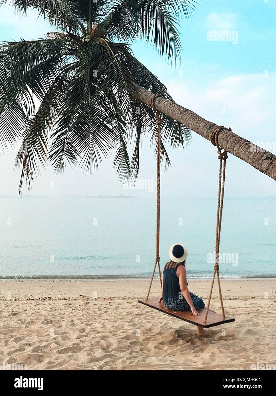 Woman sitting on swing hanging on palm tree and looking at calm blue ocean on paradise sandy beach Stock Photo