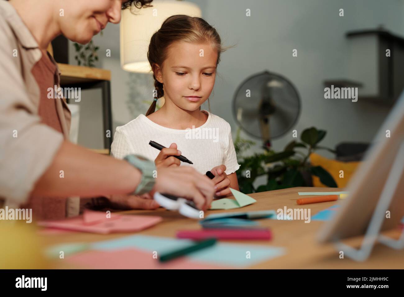 Cute girl looking at her mother folding paper while creating origami at leisure after watching online course or masterclass Stock Photo