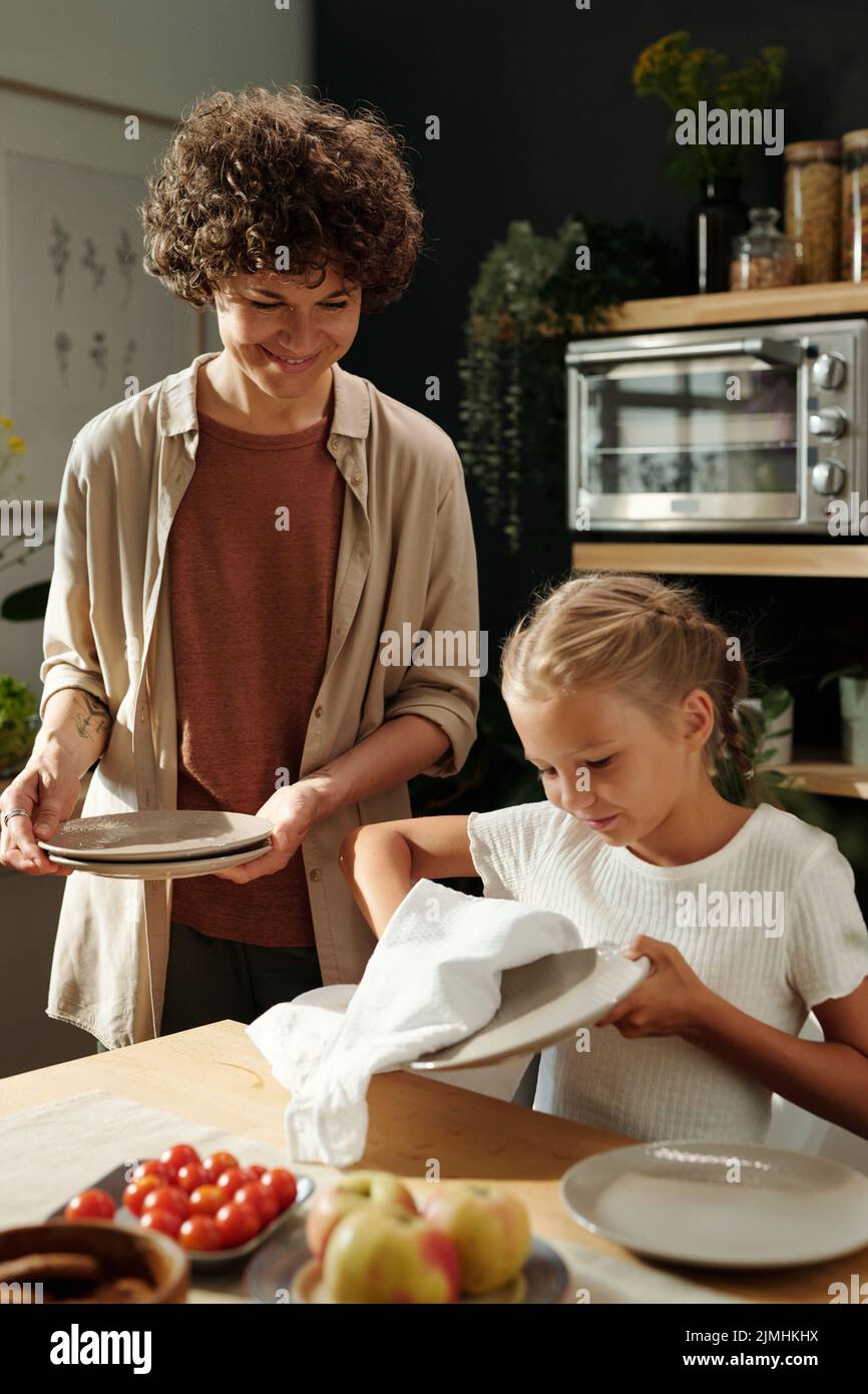 Happy young woman in casualwear looking at her youthful daughter wiping clean plates after