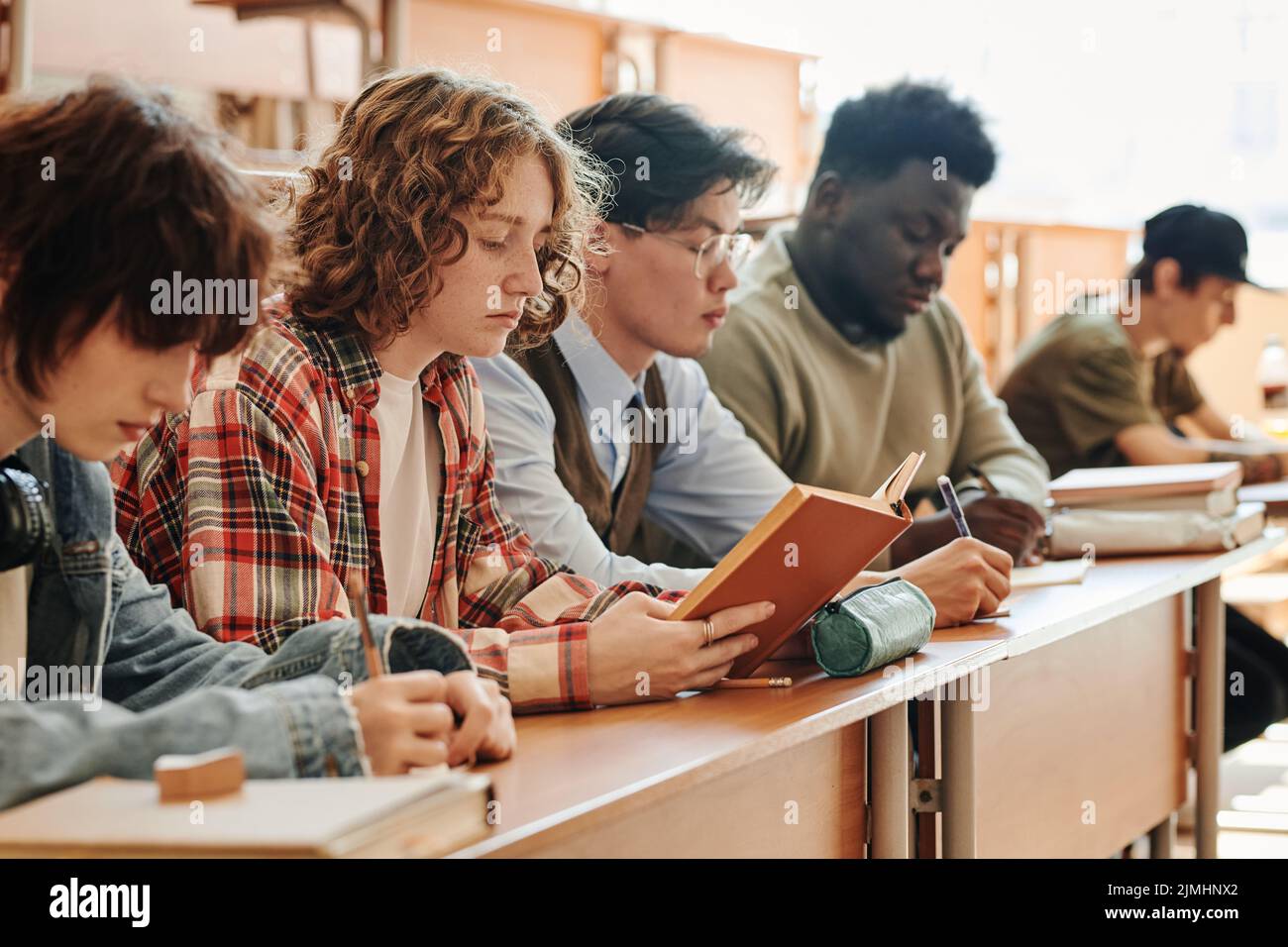 Row of serious multicultural teenage students of university sitting by desk at lecture, making notes in copybooks and reading Stock Photo