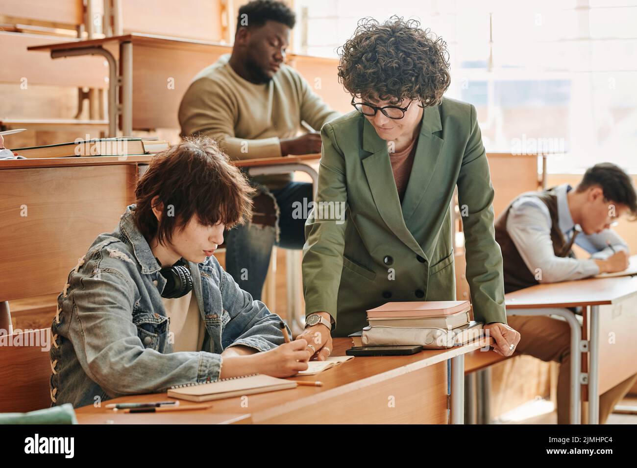 Confident teacher bending over teenage girl sitting by desk and pointing at her notes while explaining point of the task at lesson Stock Photo
