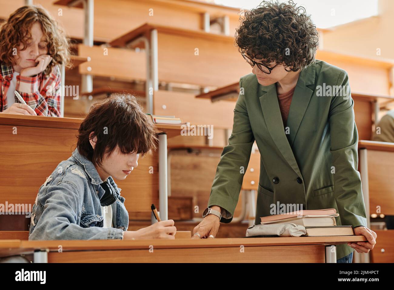 Teenage student making notes in copybook while teacher bending over her, pointing at them and checking individual written task at lesson Stock Photo
