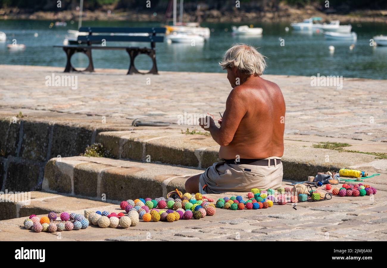 Old tanned man making handmade ball key chains with colored threads, sitting on the quay of a marina. Stock Photo